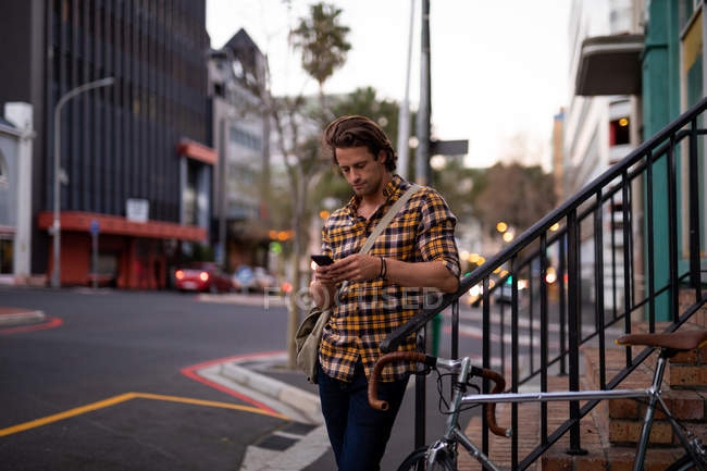 Front view of a young Caucasian man standing next to a bike and using a smartphone standing in a city street in the evening — Stock Photo