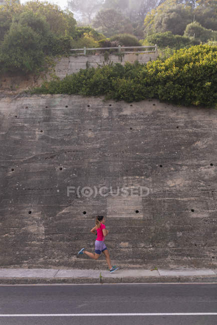 Vista laterale di una giovane donna caucasica che indossa vestiti sportivi che passano davanti a un alto muro di contenimento durante un allenamento in una giornata di sole — Foto stock