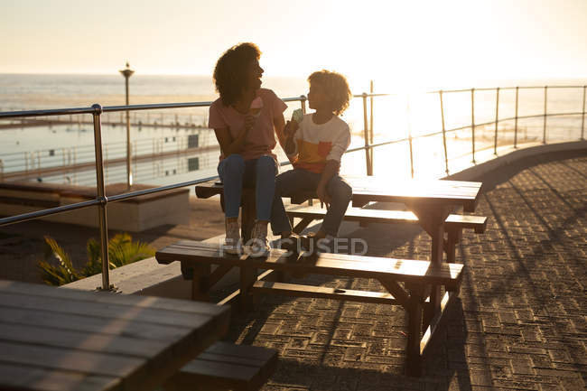 Vue de face d'une femme métissée et de son fils pré-adolescent profitant du temps passé ensemble au bord de la mer, souriant et mangeant de la crème glacée assis sur une table de pique-nique par une journée ensoleillée — Photo de stock