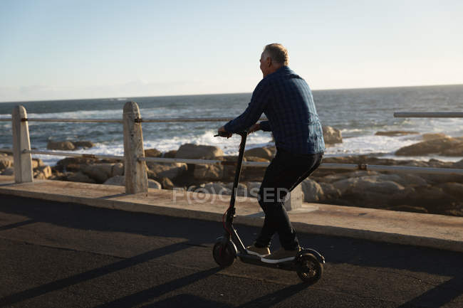 Vue arrière d'un homme caucasien mature chevauchant un scooter e par la mer au coucher du soleil — Photo de stock