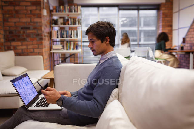 Vista laterale da vicino di un giovane caucasico seduto su un divano utilizzando un computer portatile e uno smartphone nell'area salotto di un ufficio creativo, con colleghi seduti a lavorare alle scrivanie sullo sfondo — Foto stock
