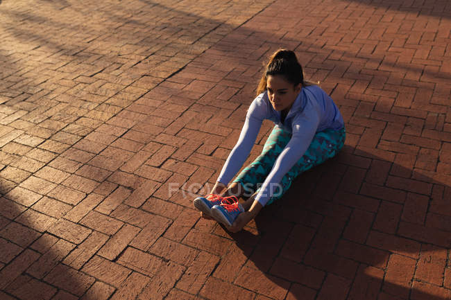 Vista elevada de una joven mujer caucásica con ropa deportiva sentada y sosteniendo sus tobillos con las piernas estiradas frente a ella mientras hace ejercicio en un parque - foto de stock