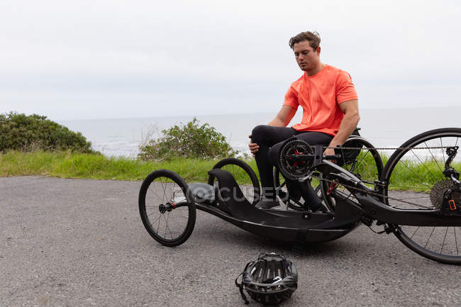 Вид спереди молодого кавказца в спортивной одежде на то, чтобы встать с инвалидного кресла на лежачий велосипед на проселочной дороге — стоковое фото