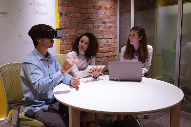 Vue de côté gros plan d'un jeune homme caucasien assis à une table portant un casque VR et levant les mains, avec une jeune femme métissée tenant un ordinateur tablette et une jeune femme caucasienne utilisant un ordinateur portable le regardant — Photo de stock