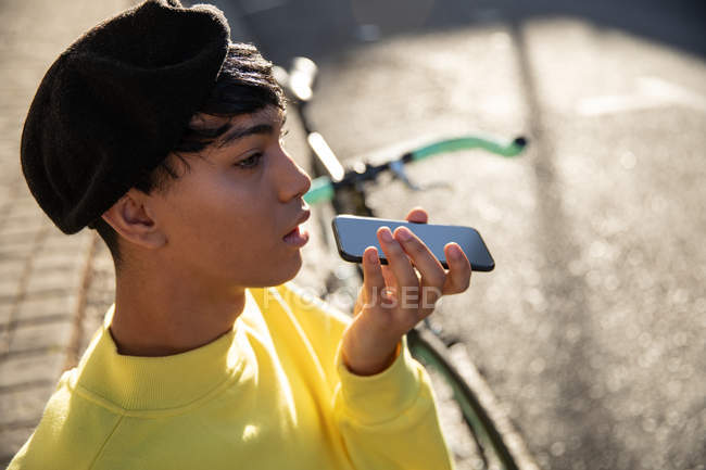 Seitenansicht eines modischen jungen Transgender-Erwachsenen mit gemischter Rasse auf der Straße, der auf dem Smartphone mit Baskenmütze und Fahrrad im Hintergrund spricht — Stockfoto