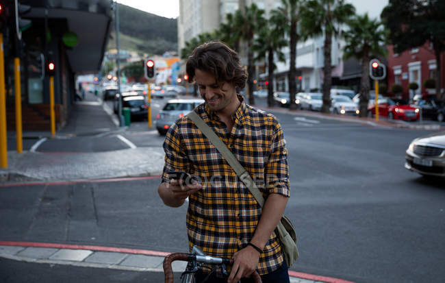Вид спереди на молодого кавказца, стоящего на городской улице, держащего велосипед и пользующегося смартфоном вечером — стоковое фото