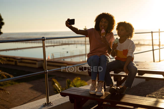 Vista frontal de una mujer de raza mixta y su hijo preadolescente disfrutando del tiempo juntos junto al mar, tomando una selfie y comiendo helado sentado en una mesa de picnic en un día soleado - foto de stock