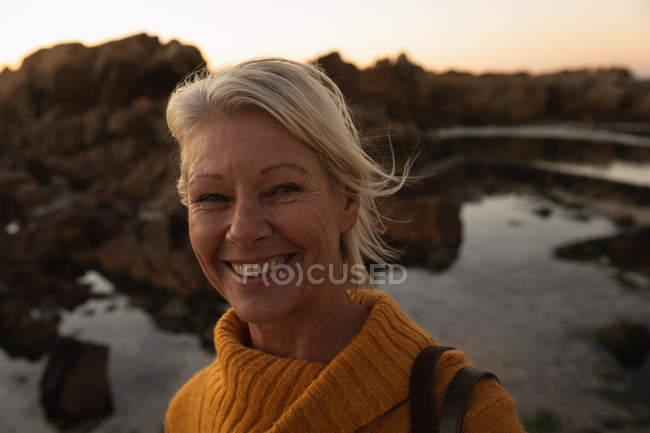 Retrato de uma mulher madura caucasiana sorrindo para a câmera junto ao mar ao pôr do sol — Fotografia de Stock