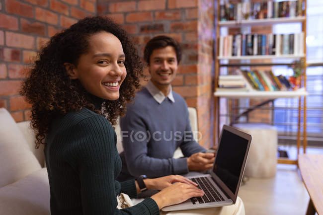 Вид сбоку: молодая женщина смешанной расы, сидящая за ноутбуком, поворачивающаяся и улыбающаяся на камеру в гостиной творческого кабинета, с молодым коллегой-кавказцем, сидящим на заднем плане, улыбающимся — стоковое фото