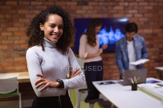 Vista frontal de cerca de una joven mujer de raza mixta que trabaja en la oficina de un negocio creativo de pie con los brazos cruzados sonriendo a la cámara, un colega masculino y femenino trabajando juntos en el fondo - foto de stock