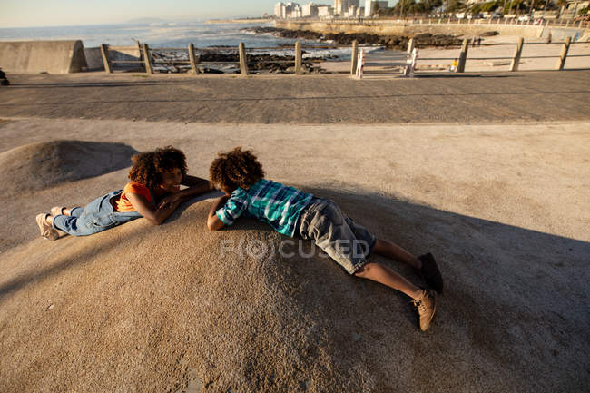 Vista lateral de una joven mestiza y su hijo preadolescente disfrutando del tiempo juntos jugando en un parque infantil junto al mar, tumbados en el suelo y mirándose en un día soleado - foto de stock