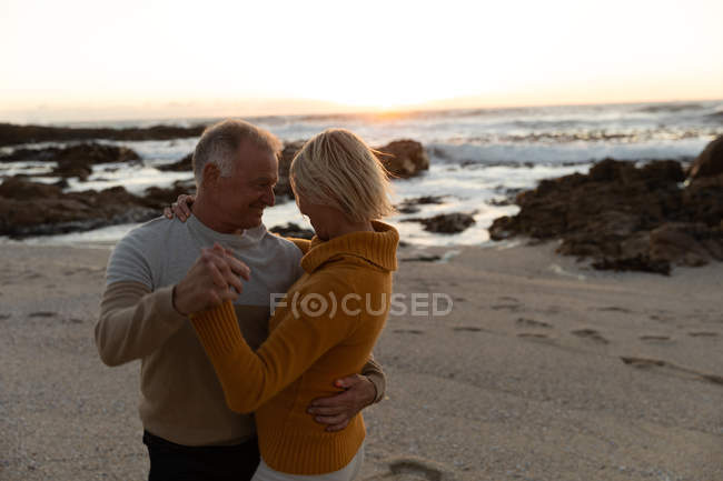 Vista lateral de un hombre caucásico maduro y una mujer bailando juntos en una playa al atardecer - foto de stock