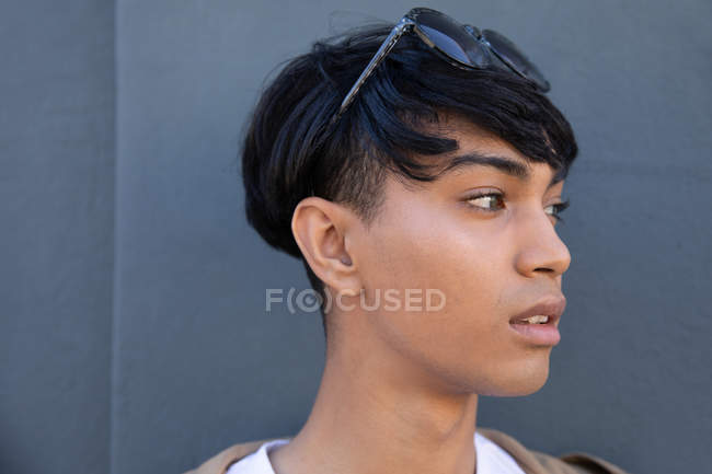 Vista lateral de un joven transgénero mestizo de moda en la calle, contra una pared gris - foto de stock