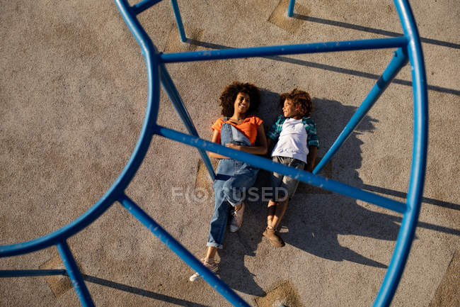 Frontansicht einer jungen Mischlingshündin und ihres vorpubertären Sohnes, die an einem sonnigen Tag zusammen auf einem Spielplatz spielen, auf dem Boden unter einem Klettergerüst liegend — Stockfoto