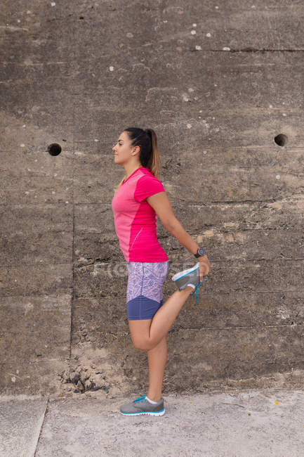Vista lateral de uma jovem caucasiana vestindo roupas esportivas em pé na frente de uma parede em uma rua, segurando o pé e esticando a perna durante um treino — Fotografia de Stock