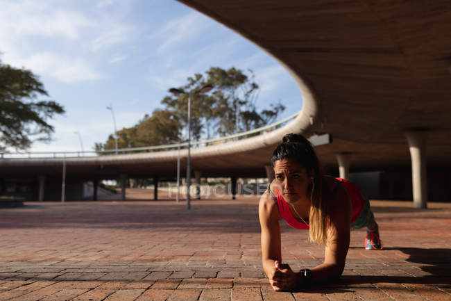 Vista frontal de uma jovem caucasiana vestindo roupas esportivas fazendo o exercício de prancha durante um treino em um dia ensolarado em um parque — Fotografia de Stock