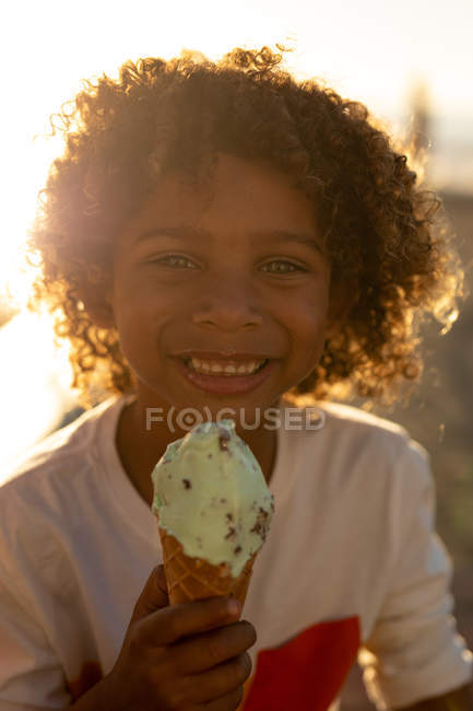 Retrato de un niño preadolescente sonriente con el pelo rizado comiendo un helado junto al mar, retroiluminado por el sol poniente - foto de stock
