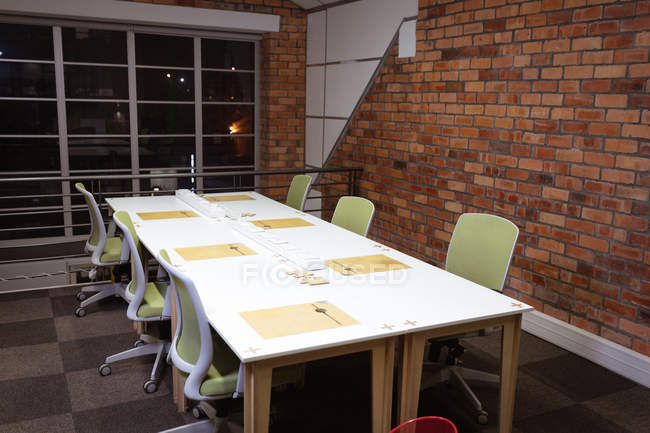 Uma mesa branca e seis cadeiras em uma sala de reuniões vazia com uma grande janela e parede de tijolo exposto no escritório de um negócio criativo — Fotografia de Stock