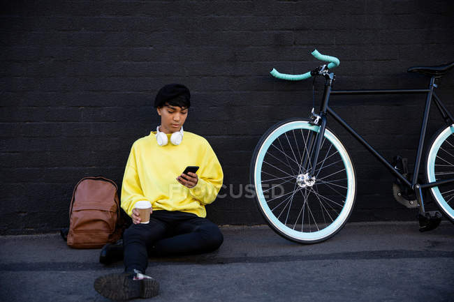 Vue de face d'un jeune transgenre de race mixte à la mode adulte dans la rue, textos sur le smartphone assis à côté d'un vélo — Photo de stock