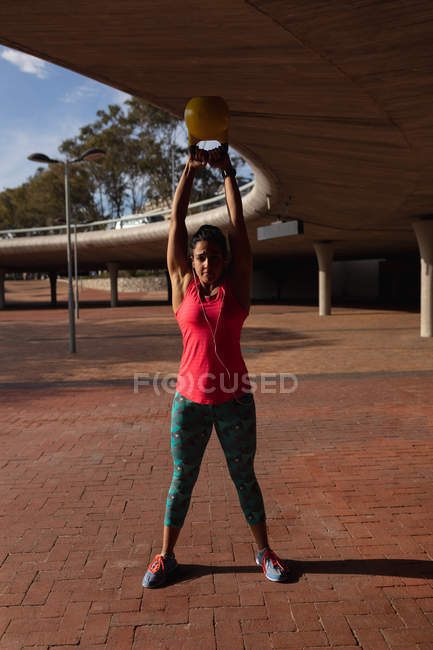 Vista frontal close-up de uma jovem caucasiana vestindo roupas esportivas segurando um peso kettlebell sobre a cabeça durante um treino em um dia ensolarado em um parque — Fotografia de Stock