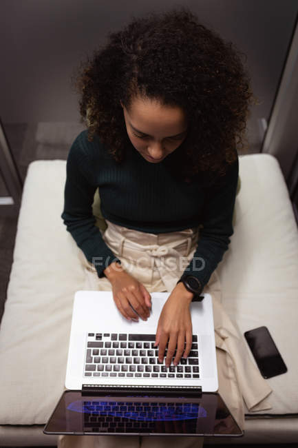 Вид спереду крупним планом молода змішана жінка, що сидить у лаунж-зоні, працює на ноутбуці в офісі креативного бізнесу, смартфон поруч з нею — стокове фото