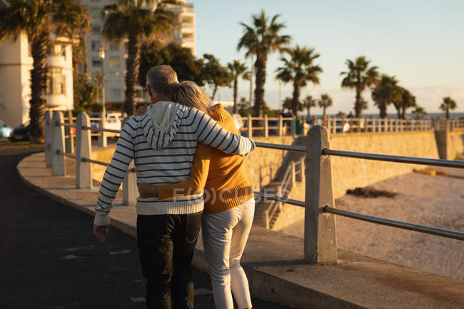 Vue arrière d'un homme et d'une femme caucasiens matures embrassant la mer au coucher du soleil, avec des palmiers et des bâtiments en arrière-plan — Photo de stock