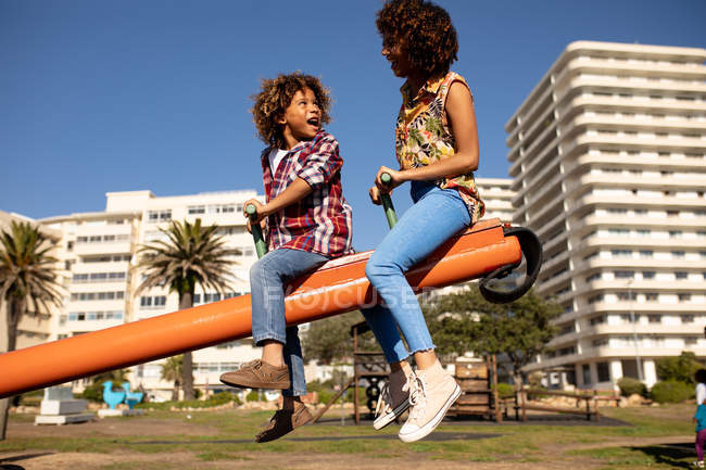 Side view of a young mixed race woman and her pre-teen son enjoying time together playing at a playground, sitting on a seesaw on a sunny day with buildings in the background — Stock Photo