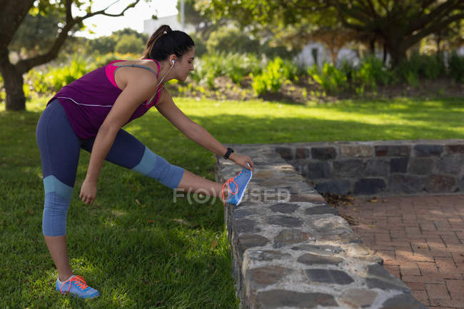 Vista lateral de uma jovem caucasiana vestindo roupas esportivas tocando seus dedos dos pés com a perna em uma parede baixa durante um treino em um parque — Fotografia de Stock
