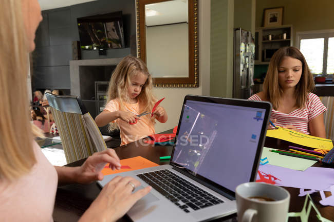 Side view close up of a young Caucasian woman using her laptop computer and doing crafts with her tween and younger daughters in their sitting room — Stock Photo