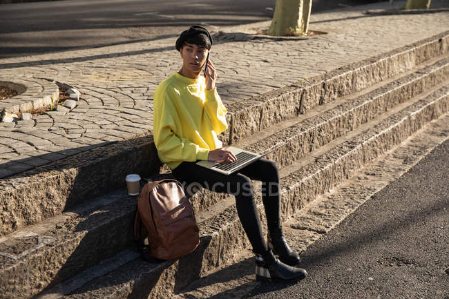 Vista frontal de cerca de un joven transgénero de raza mixta de moda en la calle, utilizando un ordenador portátil y hablando en el teléfono inteligente - foto de stock
