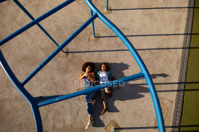 Vista frontale di una giovane donna di razza mista e del suo figlio pre-adolescente che si divertono insieme giocando in un parco giochi, sdraiati a terra sotto una cornice di arrampicata e scattando selfie in una giornata di sole — Foto stock