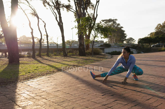 Front view of a young Caucasian woman wearing sports clothes squatting down and touching the ground with one leg stretched out while working out in a park — Stock Photo