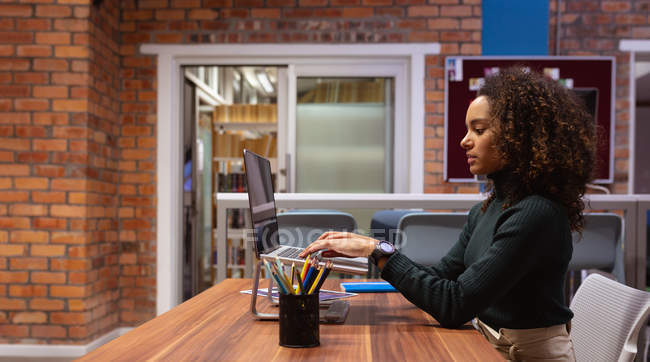 Side view close up of a young mixed race woman working in the office of a creative business sitting at a desk using a laptop computer — Stock Photo