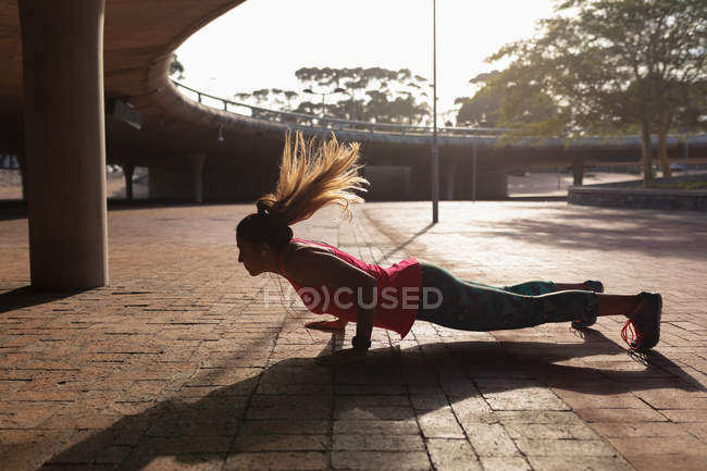 Side view of a young Caucasian woman wearing sports clothes doing push ups during a workout on a sunny day in a park, her hair flying in the air, backlit by sunlight — Stock Photo