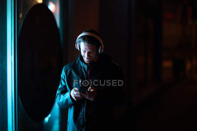 Frontansicht eines jungen kaukasischen Mannes, der nachts auf einer Straße steht, Musik mit Kopfhörern hört und auf ein Smartphone blickt — Stockfoto