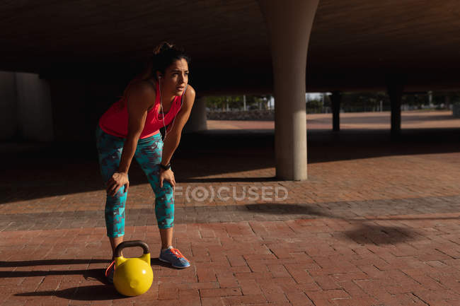 Вид сбоку на молодую белую женщину в спортивной одежде, наклоняющуюся вперед с руками на коленях, с гиревым грузом рядом с ней, отдыхающую во время тренировки в солнечный день в парке — стоковое фото