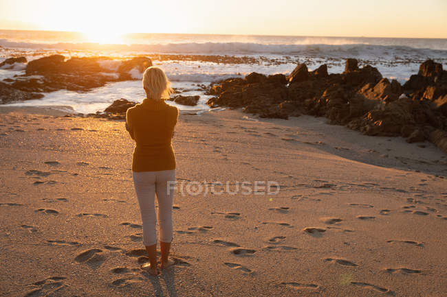 Vista trasera de una mujer caucásica madura admirando la vista junto al mar al atardecer - foto de stock