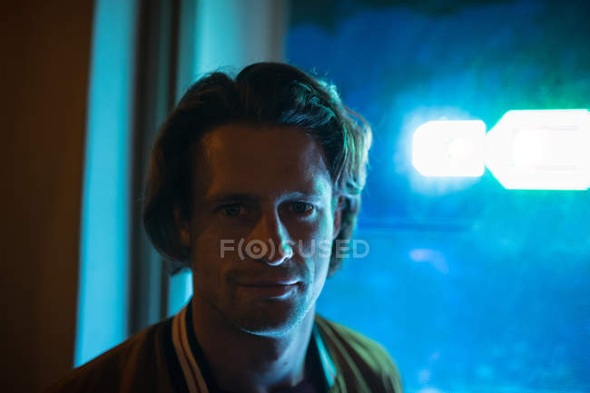 Portrait of a young Caucasian man looking to camera in the evening with blue neon light from a shop window behind him — Stock Photo