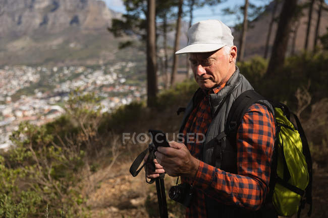 Vista lateral de un hombre caucásico maduro usando un teléfono inteligente y sosteniendo bastones nórdicos en un entorno rural - foto de stock