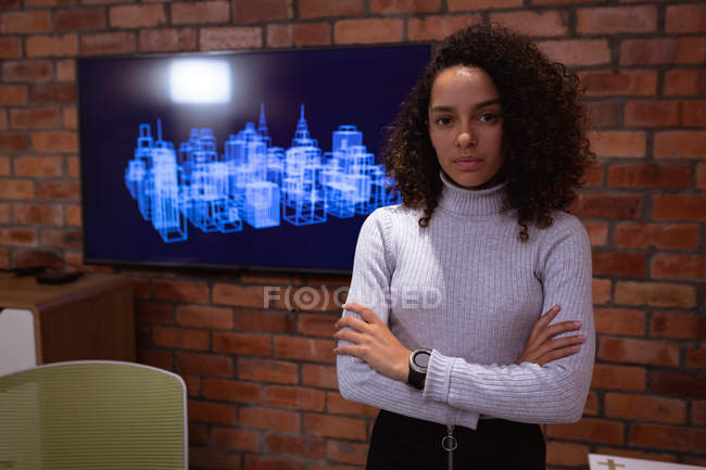 Portrait gros plan d'une jeune femme métissée debout les bras croisés regardant droit devant la caméra dans le bureau d'une entreprise créative, un écran monté sur un mur de briques exposé en arrière-plan — Photo de stock