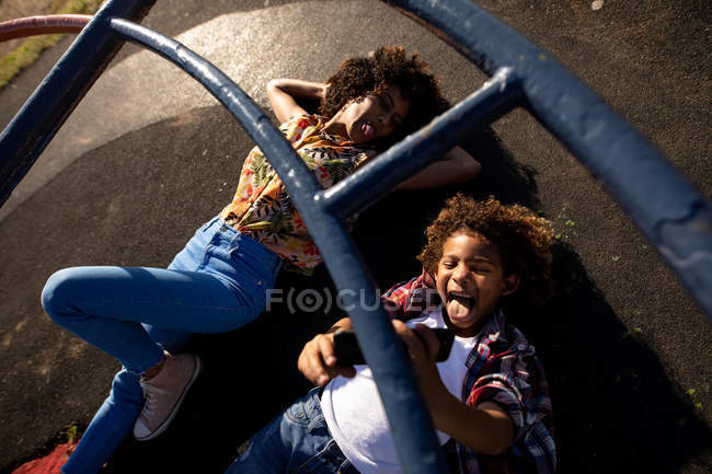 Вид спереди на молодую женщину смешанной расы и ее сына-подростка, наслаждающуюся временем вместе, играющую на детской площадке, лежащую на земле под скалолазанием и делающую селфи в солнечный день — стоковое фото