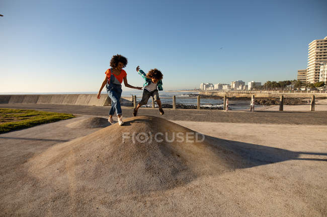 Vue latérale d'une jeune femme métissée et de son fils pré-adolescent jouissant du temps passé ensemble sur une aire de jeux au bord de la mer, le garçon sautant par une journée ensoleillée — Photo de stock