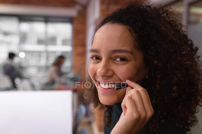 Retrato de cerca de una joven mujer de raza mixta sonriente que trabaja en la oficina de un negocio creativo con un auricular de teléfono y mirando a la cámara, con colegas trabajando en el fondo - foto de stock