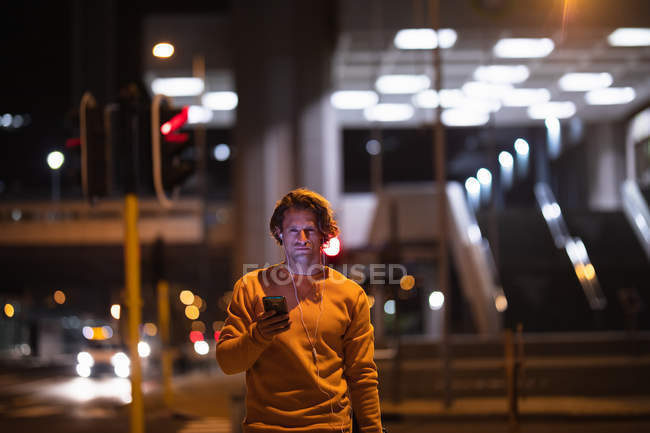 Front view of a young Caucasian man in the street at night holding a smartphone and wearing earphones on, looking to camera — Stock Photo