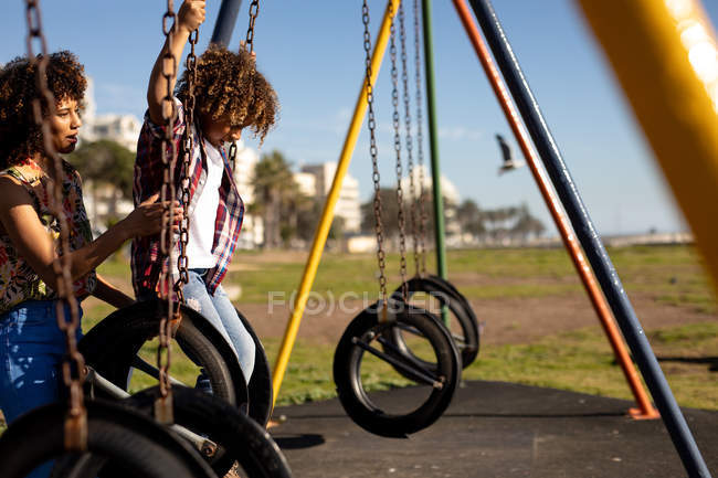 Vista laterale di una giovane donna di razza mista e del suo figlio pre-adolescente che si divertono insieme giocando in un parco giochi, oscillando in una giornata di sole — Foto stock