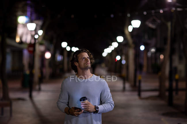 Front view of a young Caucasian man walking in a street at night holding a smartphone and a takeaway coffee and looking away — Stock Photo