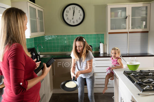 Вид спереди на молодую кавказскую беременную женщину, которая печет блинчики на кухне дома со своей младшей дочерью — стоковое фото