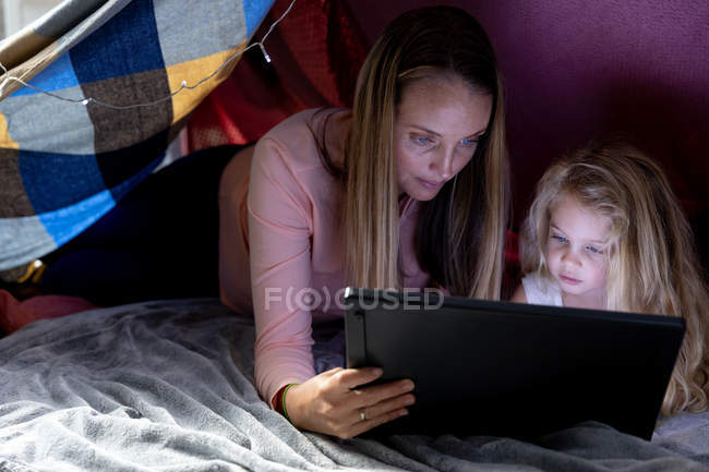 Передній погляд на молоду кавказьку жінку з її маленькою донькою, яка використовує планшет разом у наметі, виготовленому з ковдр. — стокове фото