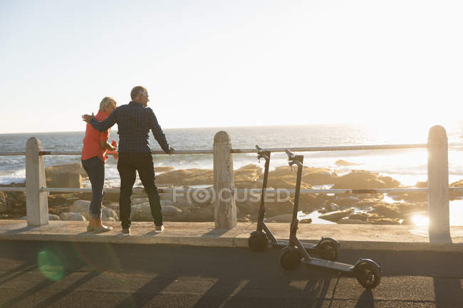 Visão traseira de um homem e uma mulher caucasianos maduros abraçando ao lado de e scooters pelo mar no por do sol — Fotografia de Stock
