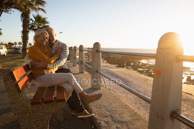 Vorderansicht eines reifen kaukasischen Mannes und einer Frau, die auf einer Bank sitzen und sich bei Sonnenuntergang am Meer umarmen, mit Palmen im Hintergrund — Stockfoto